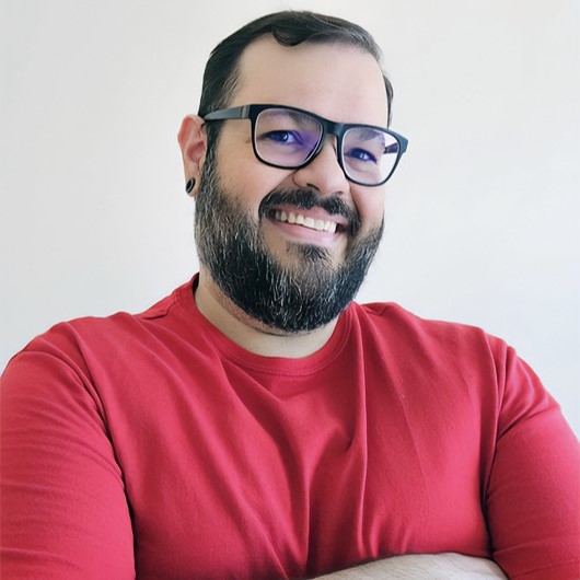 Foto de homem de pele branca, cabelos e barba curta e preta, usa óculos, sorri, veste camisa vermelha