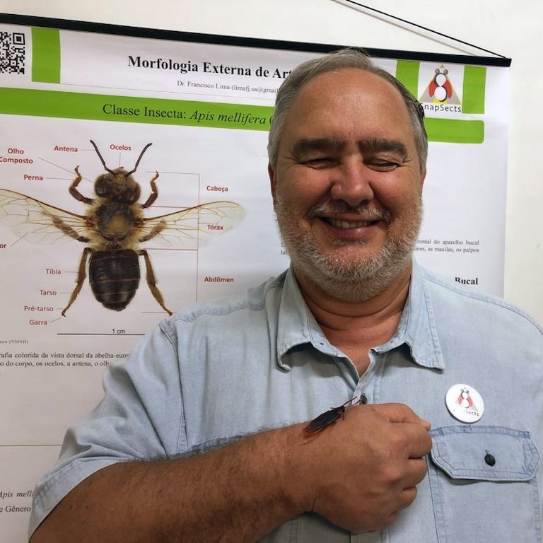 À direita da fotografia, em pé, homem segura com a mão direita um bloquinho de resina, dentro do qual está preservado um besouro. À esquerda, parcialmente oculto pelo corpo do professor, há um banner apresentando a fotografia colorida da abelha europeia, Apis mellifera. Acima do bolso esquerdo da camisa do professor, há um bottom do SnapSects.
