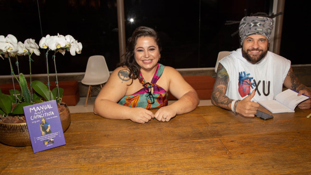 Duas pessoas cadeirantes, com os braços apoiados sobre a mesa. No jarro de flores brancas está apoiado o livro. A Carolina é uma mulher branca, de cabelos longos e lisos, tem uma tatuagem no ombro direito, veste uma roupa estampada e as alças estão cruzadas no pescoço. O Billy é um homem pardo, usa dreadlocks e uma faixa grossa, cinza e estampada e tem os braços cobertos por tatuagens. Veste camisa branca sem mangas.