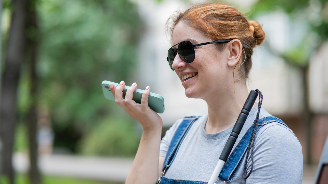 Mulher de pele branca, cabelos lisos e ruivos amarrado com coque. Usa óculos escuros, veste camisa cinza, macacãop jeans, tem apoiodo no corpo uma bengada e segura um celular com uma das mãos em direção da boca.