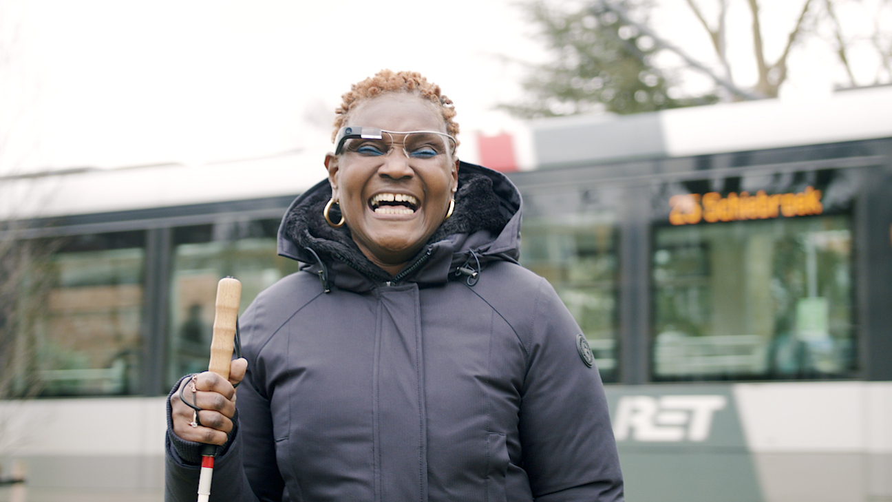 Mulher negra com cabelo crespos e curtos ruivos, casaco pret e argolhas douradas nas duas orelhas. Ela exibe um sorriso largo, segura uma bengala e um óculos da Envision.