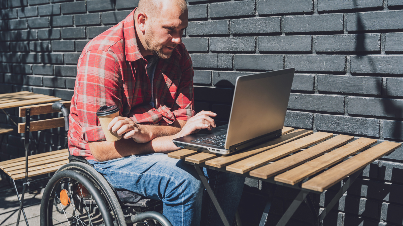 Homem careca, de pele branca, sentado em uma cadeira de rodas. Apoia uma notebook em uma mesa de madeira