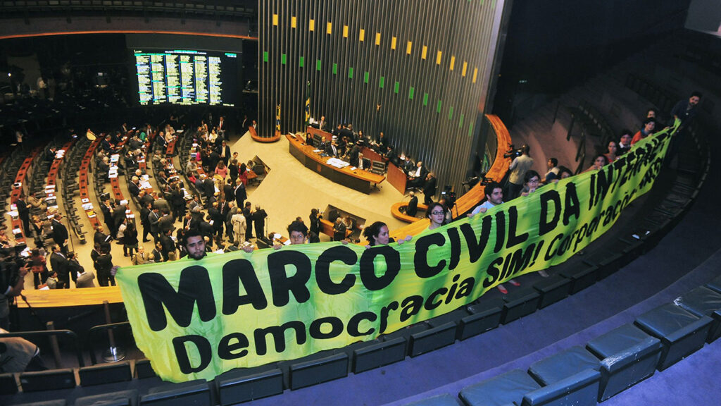 Câmara dos Deputados do Brasil. Em destaque, há um grupo de pessoas segurando uma grande faixa verde com letras pretas que diz 'MARCO CIVIL DA INTERNET - Democracia SIM! Corporações NÃO!'. Essa faixa está posicionada no andar superior do auditório, acima dos assentos dos legisladores. Ao fundo, vários parlamentares e funcionários ocupando as cadeiras e circulando pelo plenário. O ambiente é formal, com muitas cadeiras dispostas em fileiras semicirculares voltadas para uma mesa principal onde provavelmente estão posicionados os líderes ou o presidente da sessão
