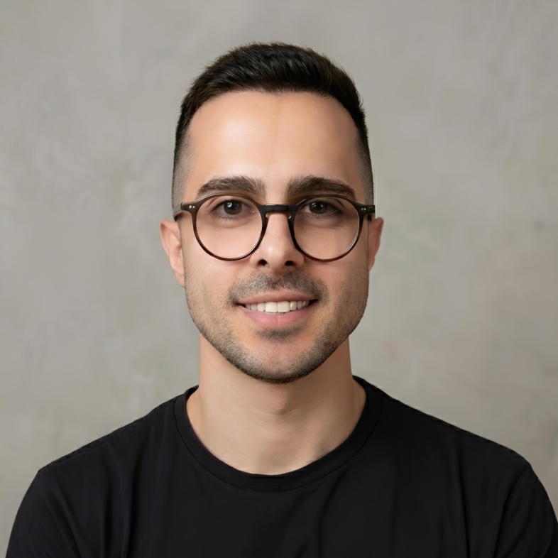 Homem enquadrado do peito para cima, tem cabelo curto, escuro e bem aparado, com laterais mais baixas. Usa óculos de armação redonda e fina, de cor escura. O rosto é limpo, com leve barba por fazer, e ele está sorrindo. Veste uma camiseta preta simples, sem estampas. O fundo é liso, em tom claro e levemente texturizado.