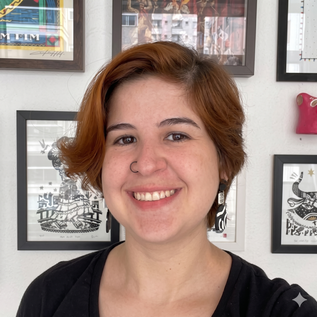 Foto em primeiro plano de uma pessoa sorrindo para a câmera. Ela tem pele clara, cabelos curtos em tom ruivo, penteados lateralmente, e usa camiseta preta, brincos compridos em preto e branco e um piercing de argola no nariz. Ao fundo, há uma parede clara decorada com diversos quadros de ilustrações, predominantemente em preto e branco, além de obras coloridas na parte superior. No canto inferior direito da imagem aparece um pequeno símbolo gráfico em forma de estrela.